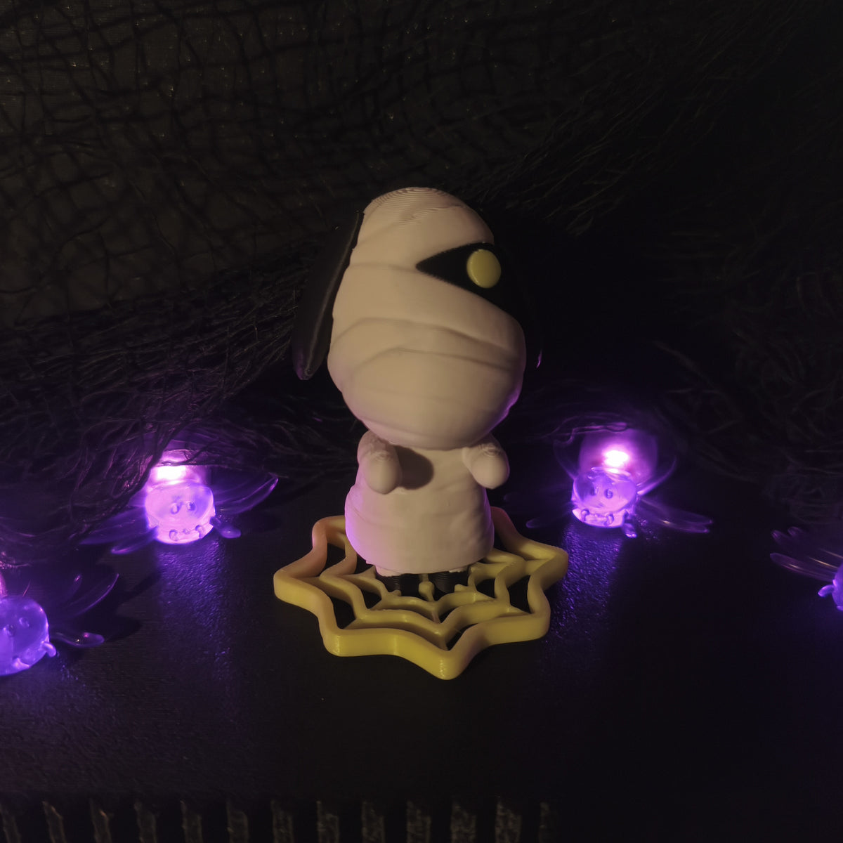 Figura Pupas impresa en 3D: momia blanca sobre base de telaraña amarilla, vista frontal